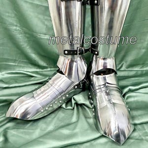 May include: A pair of silver metal shin guards with buckles. The shin guards are worn over a pair of black boots. The shin guards are made of metal plates that are joined together with rivets. The shin guards are designed to protect the shins from injury during combat.