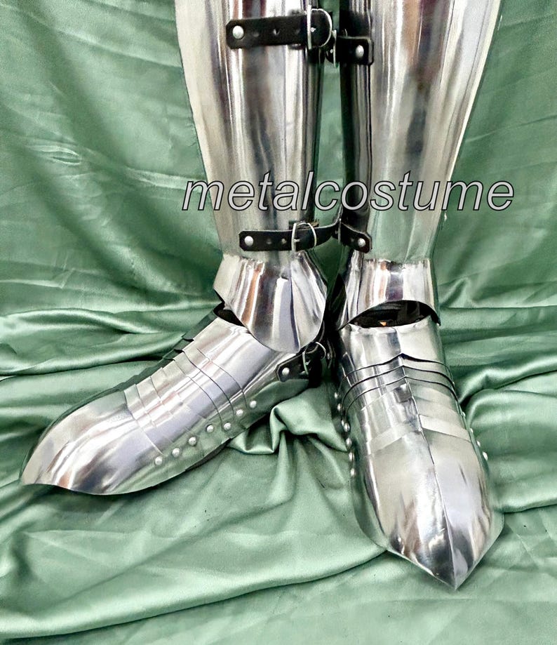 May include: A pair of silver metal shin guards with buckles. The shin guards are worn over a pair of black boots. The shin guards are made of metal plates that are joined together with rivets. The shin guards are designed to protect the shins from injury during combat.