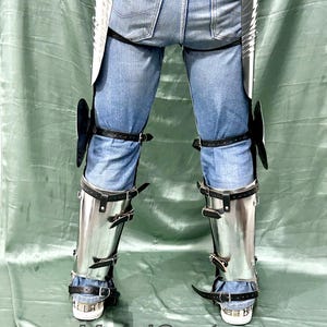 Op de afbeelding: Gepolijste metalen beenpantser met zwarte leren riemen en gespen, gedragen over een blauwe spijkerbroek. De bepantsering omvat knie- en scheenbescherming. De tekst "MetalCostume" is onderaan zichtbaar.