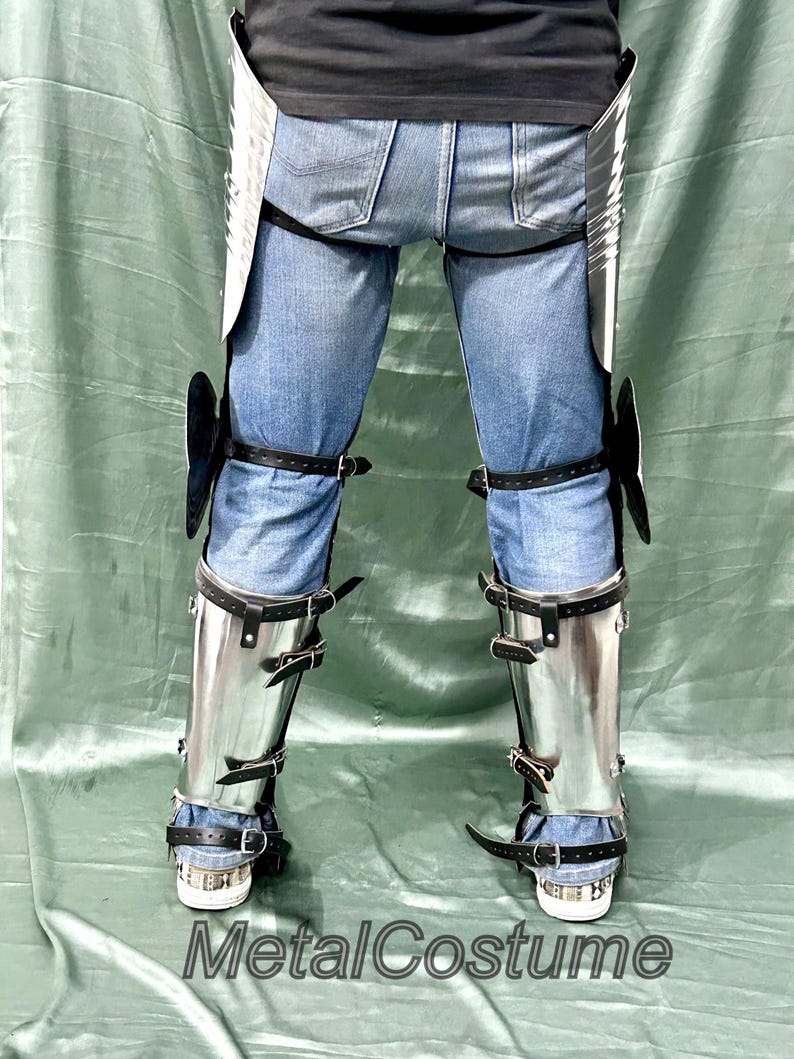Op de afbeelding: Gepolijste metalen beenpantser met zwarte leren riemen en gespen, gedragen over een blauwe spijkerbroek. De bepantsering omvat knie- en scheenbescherming. De tekst "MetalCostume" is onderaan zichtbaar.