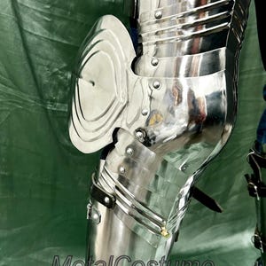 Op de afbeelding: Close-up van gepolijst, zilverkleurig metalen harnas, met gearticuleerde platen en klinknagels. Het harnas omvat een kniebeschermer en beenbescherming, die de omgeving weerspiegelt. De tekst "MetalCostume" is zichtbaar.