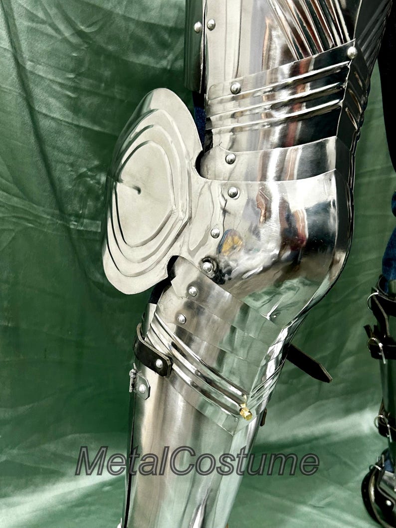 Op de afbeelding: Close-up van gepolijst, zilverkleurig metalen harnas, met gearticuleerde platen en klinknagels. Het harnas omvat een kniebeschermer en beenbescherming, die de omgeving weerspiegelt. De tekst "MetalCostume" is zichtbaar.
