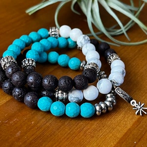 Può includere: Tre braccialetti sovrapposti con perline turchesi, bianche e nere. I braccialetti hanno dettagli argentati e un ciondolo a stella argentato.