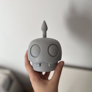May include: A gray, 3D printed figurine of a cartoon character with a single, pointed antenna on top of its head. The character has two large, round eyes and a small, toothy mouth.