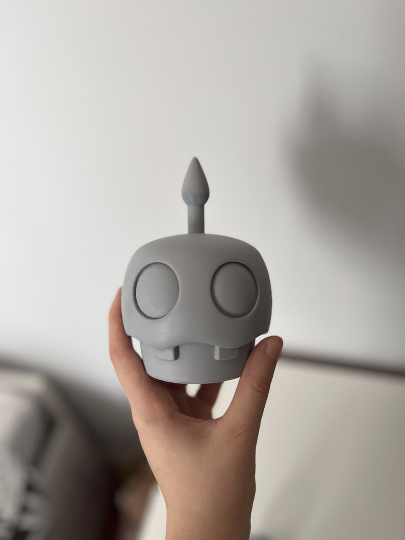 May include: A gray, 3D printed figurine of a cartoon character with a single, pointed antenna on top of its head. The character has two large, round eyes and a small, toothy mouth.
