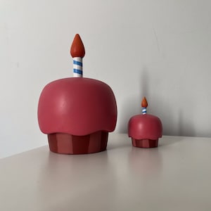 May include: Two pink and red cupcake figurines with candles. The larger cupcake has a blue and white striped candle, and the smaller cupcake has a red and white striped candle.
