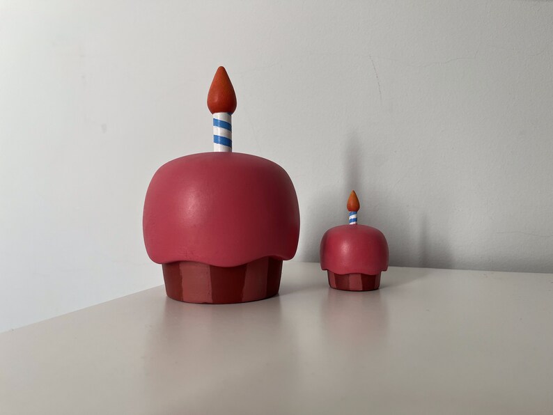 May include: Two pink and red cupcake figurines with candles. The larger cupcake has a blue and white striped candle, and the smaller cupcake has a red and white striped candle.