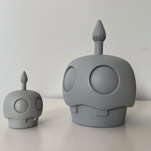 May include: Two gray ceramic figurines shaped like robots with rounded heads and single eyes. The larger figurine is taller than the smaller figurine.