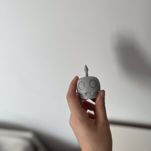 May include: A gray 3D printed figurine of a robot with a single antenna on top of its head. The robot has two large round eyes and a small mouth.