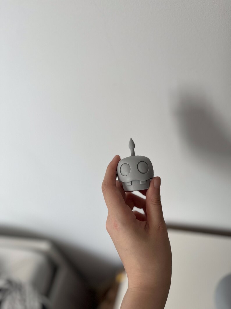 May include: A gray 3D printed figurine of a robot with a single antenna on top of its head. The robot has two large round eyes and a small mouth.