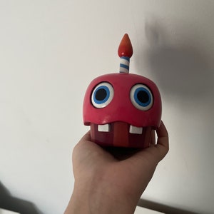 May include: A red and white cupcake-shaped animatronic toy with blue eyes and a single lit candle on top.