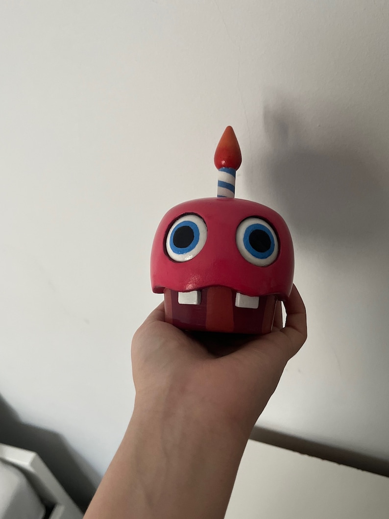 May include: A red and white cupcake-shaped animatronic toy with blue eyes and a single lit candle on top.
