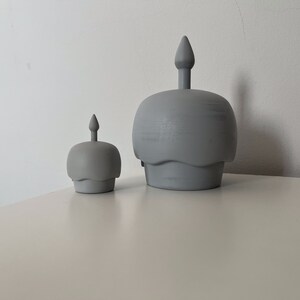 May include: Two gray ceramic candle holders shaped like cupcakes with a single candle on top of each. The larger candle holder is in the background and the smaller candle holder is in the foreground.