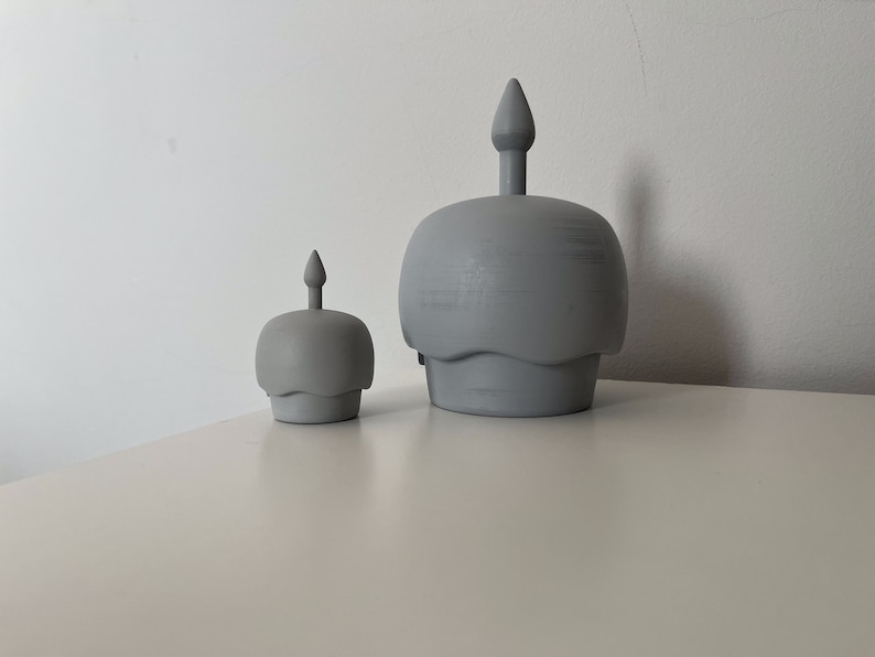 May include: Two gray ceramic candle holders shaped like cupcakes with a single candle on top of each. The larger candle holder is in the background and the smaller candle holder is in the foreground.