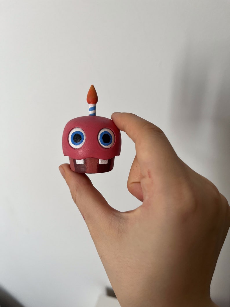May include: A red and white cupcake character with blue eyes and a single candle on top. The candle is orange and white striped.