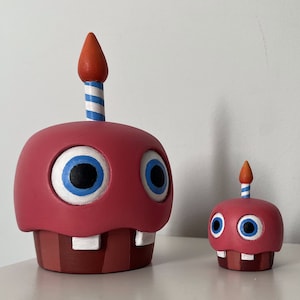 May include: Two red and white cupcake-shaped figurines with blue eyes and a single candle on top. The larger figurine has a red and white striped candle, while the smaller figurine has a blue and white striped candle.