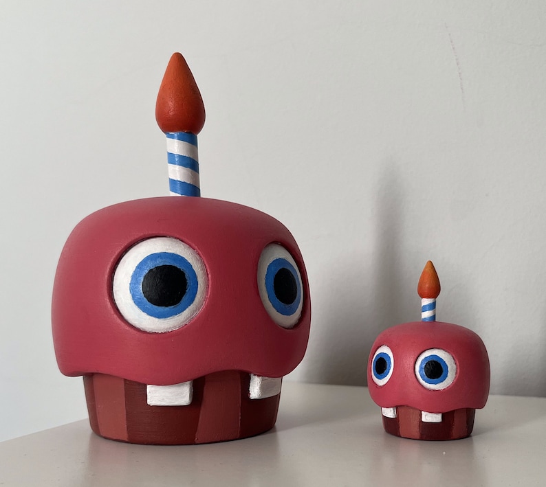 May include: Two red and white cupcake-shaped figurines with blue eyes and a single candle on top. The larger figurine has a red and white striped candle, while the smaller figurine has a blue and white striped candle.
