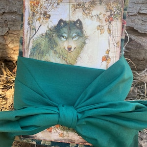 May include: A handmade journal wrapped with a teal green fabric bow. The journal cover features a detailed illustration of a wolf with green and brown fur, set against a backdrop of autumn foliage and a vintage map.