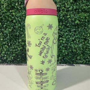 Puede incluir: Una botella de agua Owala verde con tapa y correa rojas. La botella presenta un dise&ntilde;o de dibujos animados del Grinch con el texto "You're a Mean One, Mr. Grinch!" y otros gr&aacute;ficos tem&aacute;ticos navide&ntilde;os. La botella est&aacute; dise&ntilde;ada para bebidas fr&iacute;as.