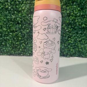 Puede incluir: Una botella de agua aislada rosa con una tapa amarilla y color melocotón. La botella presenta un dibujo lineal negro de un personaje de dibujos animados con corazones, nubes y postres. La marca "owala" está impresa en el lateral.