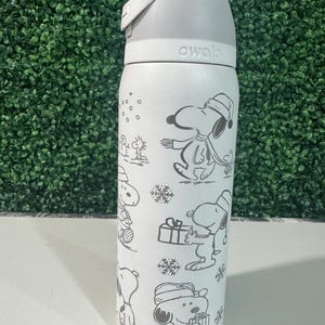 Puede incluir: Una botella de agua Owala blanca con tapa gris y un dise&ntilde;o envolvente de Snoopy con ropa de invierno. El dise&ntilde;o incluye copos de nieve y Snoopy sosteniendo regalos. La botella tiene un asa gris y un cuerpo blanco.