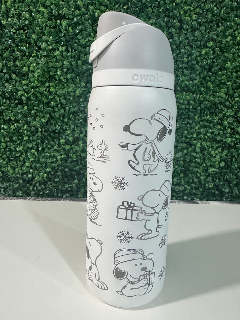 Puede incluir: Una botella de agua Owala blanca con tapa gris y un dise&ntilde;o envolvente de Snoopy con ropa de invierno. El dise&ntilde;o incluye copos de nieve y Snoopy sosteniendo regalos. La botella tiene un asa gris y un cuerpo blanco.
