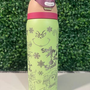 Puede incluir: Una botella de agua aislada verde con un dise&ntilde;o de dibujos animados del Grinch, copos de nieve y texto. La tapa es rosa y beige, con un bot&oacute;n y las palabras "Lift to Unlock".