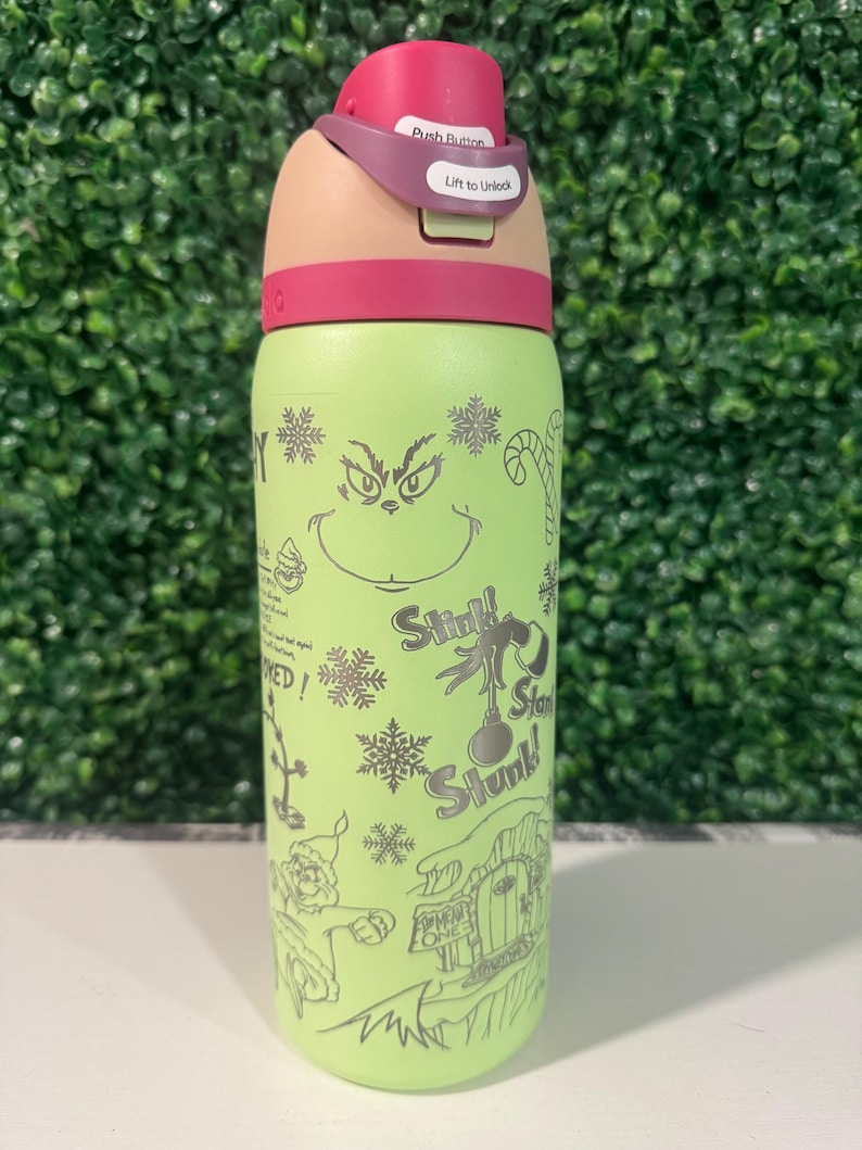 Puede incluir: Una botella de agua aislada verde con un dise&ntilde;o de dibujos animados del Grinch, copos de nieve y texto. La tapa es rosa y beige, con un bot&oacute;n y las palabras "Lift to Unlock".