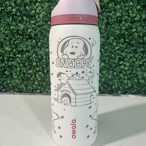 May include: A white Owala water bottle featuring a Snoopy design. The bottle has a pink and yellow lid, a maroon band, and a cartoon illustration of Snoopy and his doghouse. The text "SNOOPY" is printed below the Snoopy graphic.