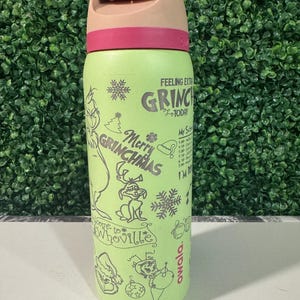 Puede incluir: Una botella de agua Owala verde claro con una tapa rosa y beige. La botella presenta ilustraciones y texto con tem&aacute;tica de Grinch, incluyendo "Merry Grinchmas" y "Feeling Extra Grinchy Today."
