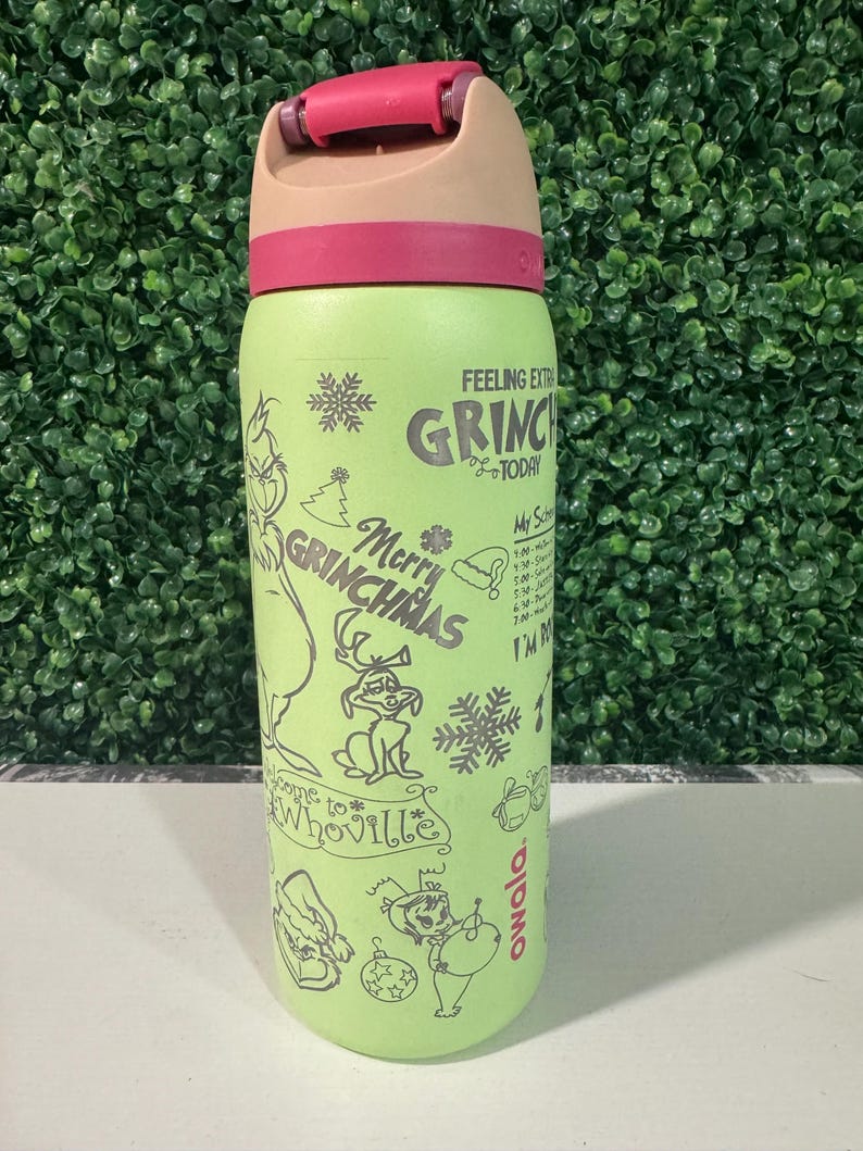 Puede incluir: Una botella de agua Owala verde claro con una tapa rosa y beige. La botella presenta ilustraciones y texto con tem&aacute;tica de Grinch, incluyendo "Merry Grinchmas" y "Feeling Extra Grinchy Today."