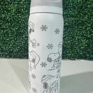 Puede incluir: Una botella de agua aislada blanca con asa y tapa grises. La botella presenta un patr&oacute;n repetido de Snoopy con ropa de invierno y copos de nieve. La botella est&aacute; dise&ntilde;ada para mantener las bebidas fr&iacute;as o calientes.