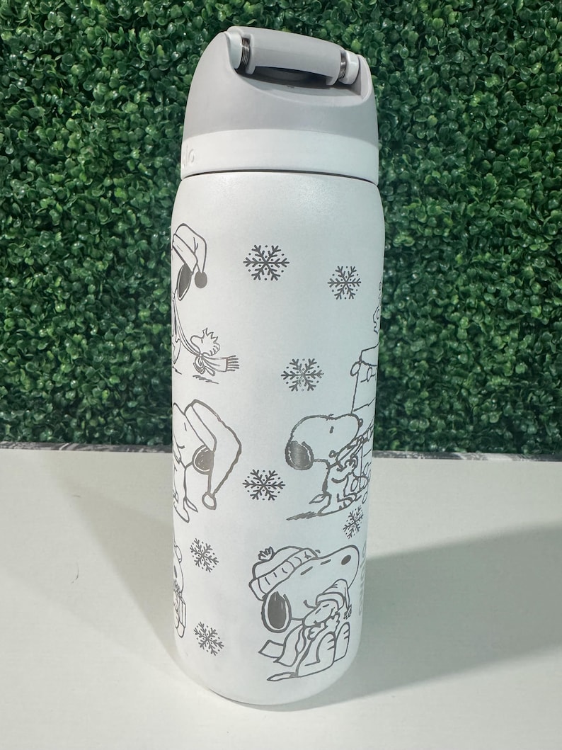 Puede incluir: Una botella de agua aislada blanca con asa y tapa grises. La botella presenta un patr&oacute;n repetido de Snoopy con ropa de invierno y copos de nieve. La botella est&aacute; dise&ntilde;ada para mantener las bebidas fr&iacute;as o calientes.