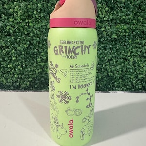 Puede incluir: Una botella de agua Owala verde con tapa rosa y beige. La botella presenta el texto "Feeling Extra Grinchy Today" y "My Schedule" con ilustraciones tem&aacute;ticas de Grinch. Dise&ntilde;ada para bebidas fr&iacute;as.