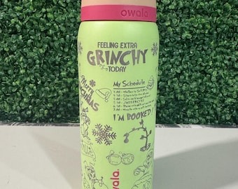 Puede incluir: Una botella de agua Owala verde con tapa rosa y beige. La botella presenta el texto "Feeling Extra Grinchy Today" y "My Schedule" con ilustraciones temáticas de Grinch. Diseñada para bebidas frías.