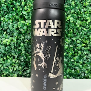 Puede incluir: Una botella de agua Owala de color negro mate con el logotipo de Star Wars en plata en la parte superior. El cuerpo muestra un gráfico de Boba Fett y Luke Skywalker con un sable de luz, rodeado de pequeñas estrellas y símbolos, con la marca Owala.