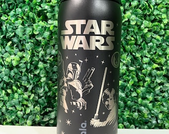 Puede incluir: Una botella de agua Owala de color negro mate con el logotipo de Star Wars en plata en la parte superior. El cuerpo muestra un gráfico de Boba Fett y Luke Skywalker con un sable de luz, rodeado de pequeñas estrellas y símbolos, con la marca Owala.