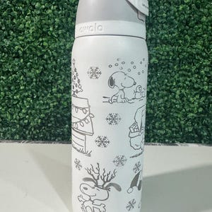 Puede incluir: Botella de agua aislada blanca con tapa y asa grises. La botella presenta una ilustraci&oacute;n en blanco y negro de Snoopy y otros personajes de Peanuts en una escena invernal, incluyendo un &aacute;rbol de Navidad y copos de nieve. La marca "awala" est&aacute; impresa en la botella.