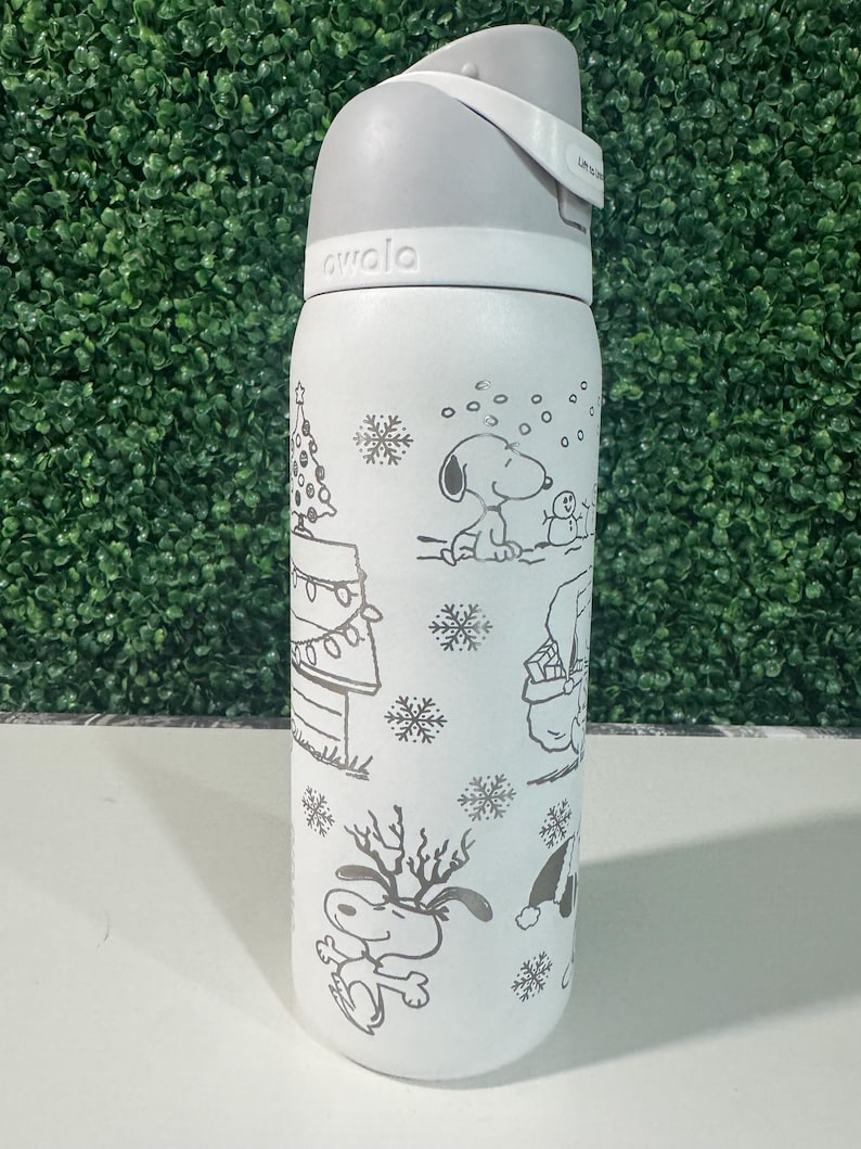 Puede incluir: Botella de agua aislada blanca con tapa y asa grises. La botella presenta una ilustraci&oacute;n en blanco y negro de Snoopy y otros personajes de Peanuts en una escena invernal, incluyendo un &aacute;rbol de Navidad y copos de nieve. La marca "awala" est&aacute; impresa en la botella.
