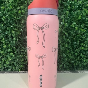 Puede incluir: Una botella de agua rosa Owala con tapa roja y gris. La botella está decorada con un patrón de lazos grises. El logotipo de Owala está impreso en la botella. La botella está diseñada para la hidratación.