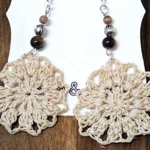 May include: A pair of handmade earrings featuring beige crocheted flower pendants. Each earring has a silver-tone hook, with a series of beads in brown and silver. The earrings are displayed on a white card against a wooden background.
