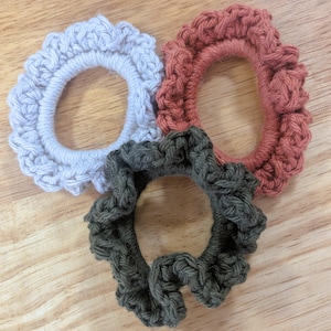 May include: Three circular hair scrunchies featuring a crocheted yarn exterior. One scrunchie is light gray, one is a muted terracotta red, and one is a dark olive green. Each accessory has a ruffled, textured edge and a flexible inner band.