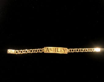 May include: A gold chain bracelet with a rectangular nameplate that reads "ASHLEY".