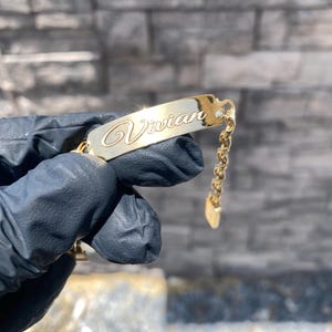 May include: A gold-colored bracelet with the name "Vivian" engraved in cursive script. The bracelet features a rectangular nameplate and a delicate chain with a heart-shaped charm. The jewelry is held against a blurred background.