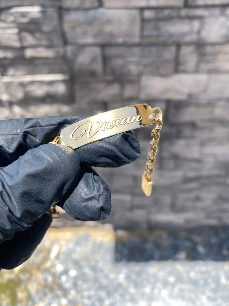 May include: A gold-colored bracelet with the name "Vivian" engraved in cursive script. The bracelet features a rectangular nameplate and a delicate chain with a heart-shaped charm. The jewelry is held against a blurred background.