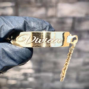 May include: A gold-colored nameplate pendant with the name "Vivian" in elegant cursive script. The pendant is rectangular with rounded edges and is attached to a delicate chain. The background is a textured gray surface.