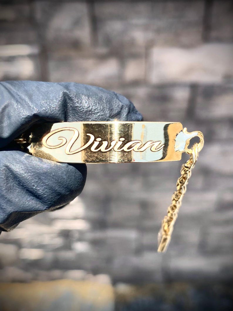 May include: A gold-colored nameplate pendant with the name "Vivian" in elegant cursive script. The pendant is rectangular with rounded edges and is attached to a delicate chain. The background is a textured gray surface.