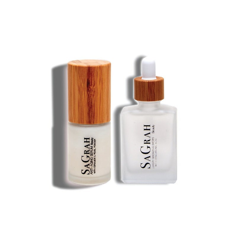 K&ouml;nnte beinhalten: Zwei Glasflaschen mit Bambusverschl&uuml;ssen, die Hautpflegeprodukte enthalten. Die Flasche links ist mit "SAGRAH ANTI-AGING SERUM Beauty WITH CERAMIDE & PEARL POWDER" beschriftet. Die Flasche rechts ist mit "SAGRAH LUKUR BRONZING DROPS Beauty WITH HYALURONIC ACID" beschriftet.