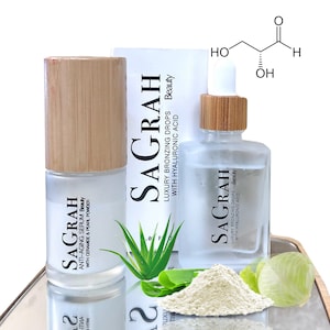 K&ouml;nnte beinhalten: Zwei Glasflaschen mit Bambusdeckeln und Etiketten mit der Aufschrift "SAGRAH" und "LUXURY BRONZING DROPS WITH HYALURONIC ACID Beauty". Die Flaschen stehen auf einer reflektierenden Oberfl&auml;che mit wei&szlig;em Pulver und gr&uuml;nen Aloe-Vera-Bl&auml;ttern.