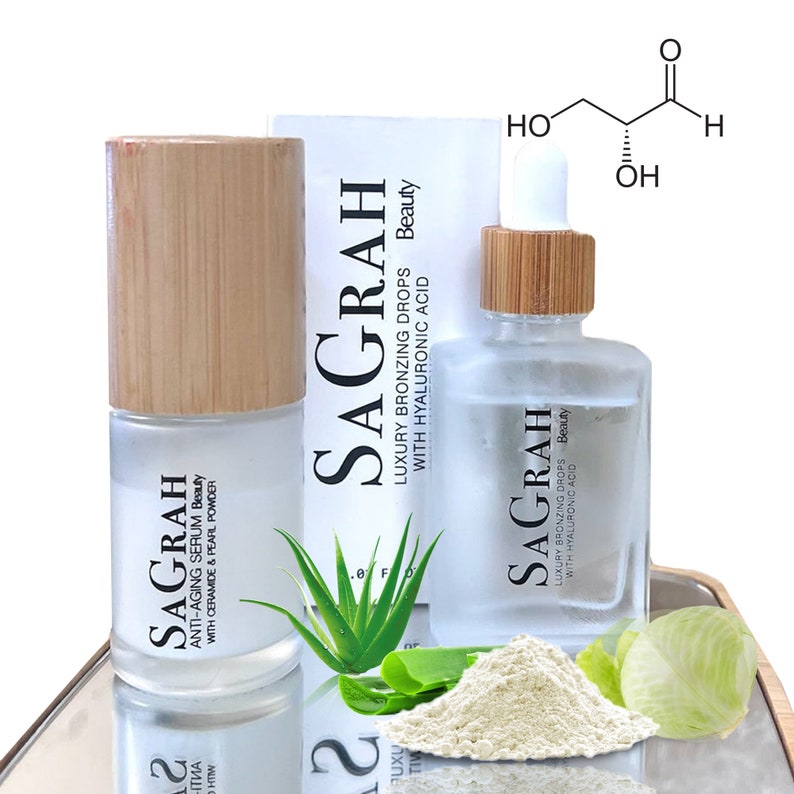 K&ouml;nnte beinhalten: Zwei Glasflaschen mit Bambusdeckeln und Etiketten mit der Aufschrift "SAGRAH" und "LUXURY BRONZING DROPS WITH HYALURONIC ACID Beauty". Die Flaschen stehen auf einer reflektierenden Oberfl&auml;che mit wei&szlig;em Pulver und gr&uuml;nen Aloe-Vera-Bl&auml;ttern.